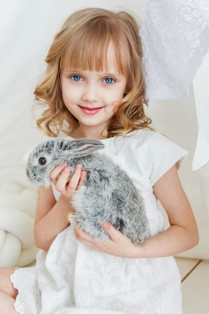 Charming young girl smiling while holding a fluffy grey bunny, radiating joy and innocence indoors.