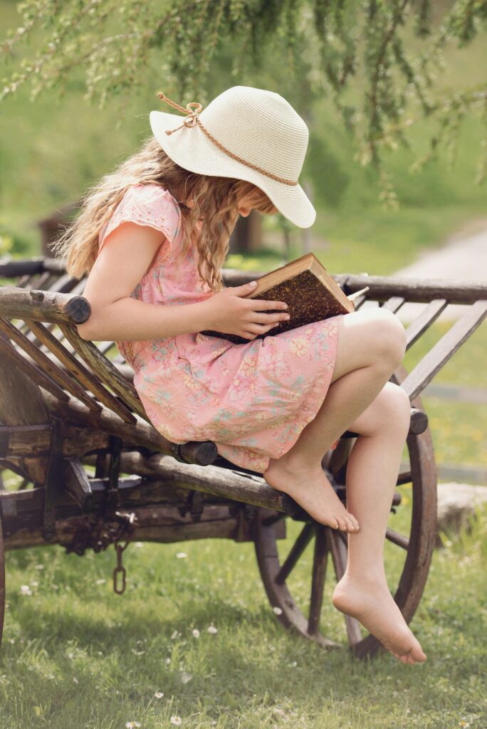 pexels-photo-261887-261887 A young girl in a hat reads a book while sitting on a rustic wooden wagon outdoors.