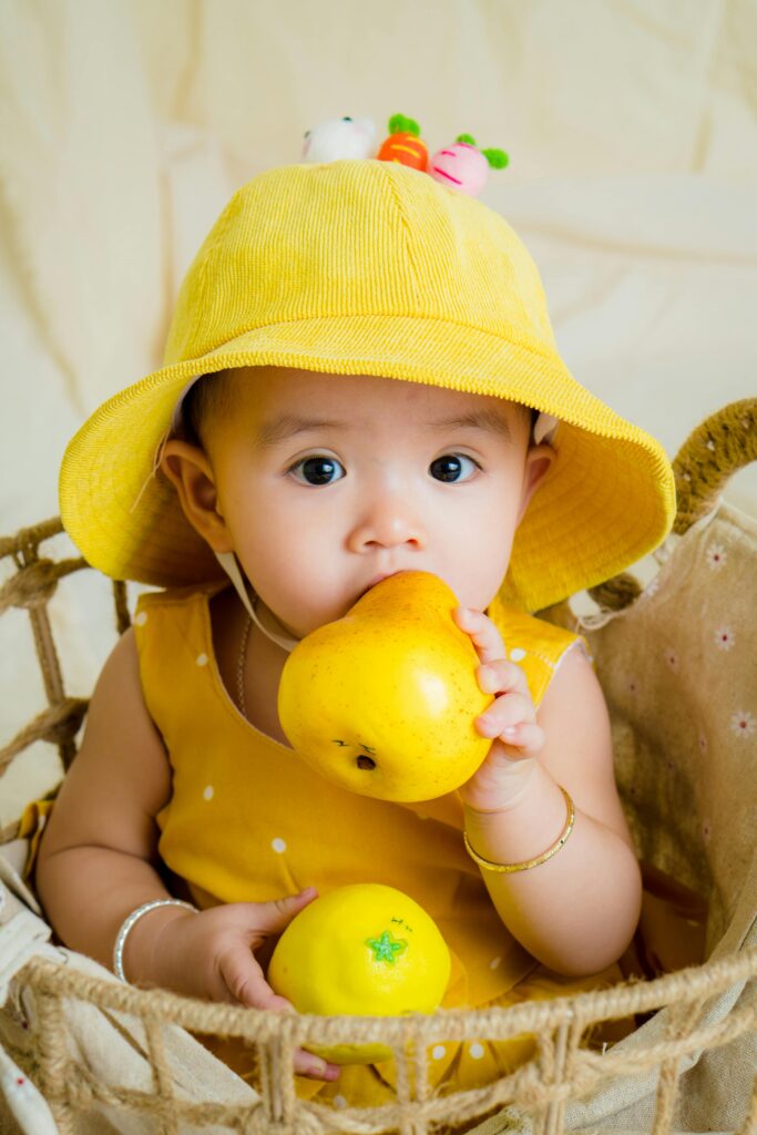 Cute baby in a yellow hat holding a pear, sitting in a basket with a playful expression.