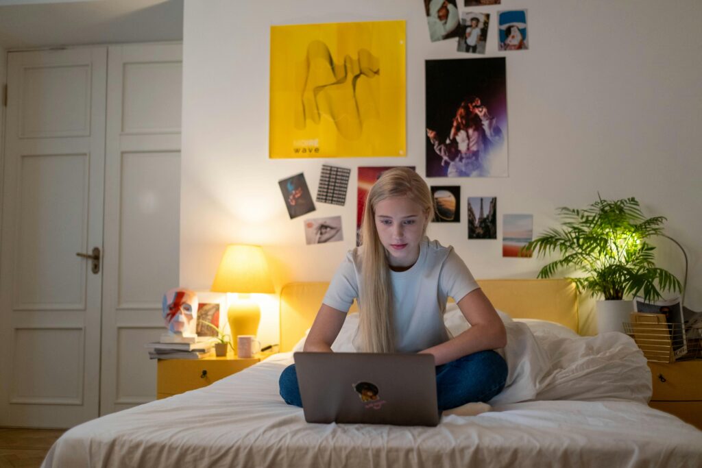 Teen girl using a laptop in her cozy bedroom lit with a warm glow, perfect for remote work or study.