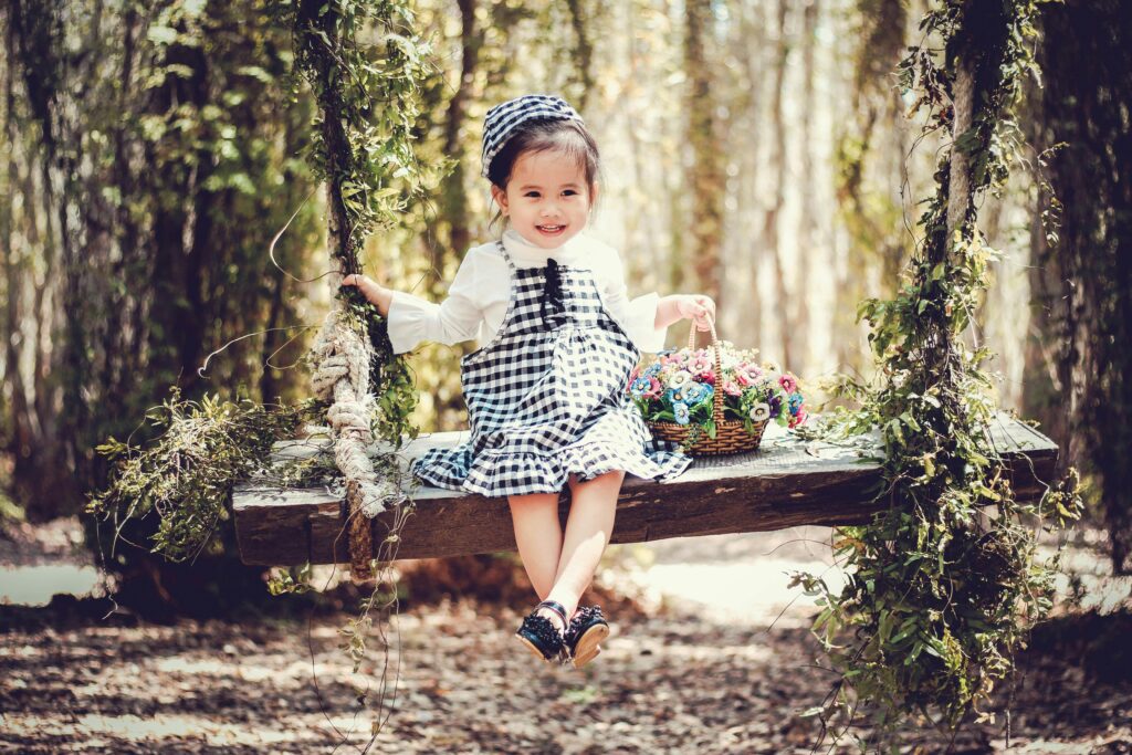Adorable child smiling on a wooden swing with a basket of flowers in a sunny park.
